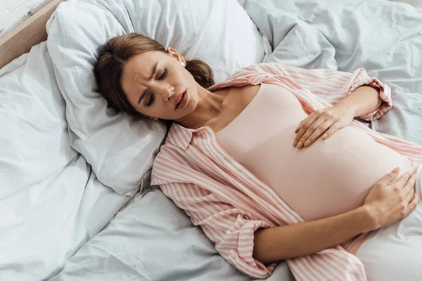 Vista aérea de la mujer embarazada emocional acostada en la cama con mueca y tocando el vientre - foto de stock