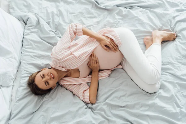 Overhead view of pregnant woman lying in bed with grimace and touching belly — Stock Photo