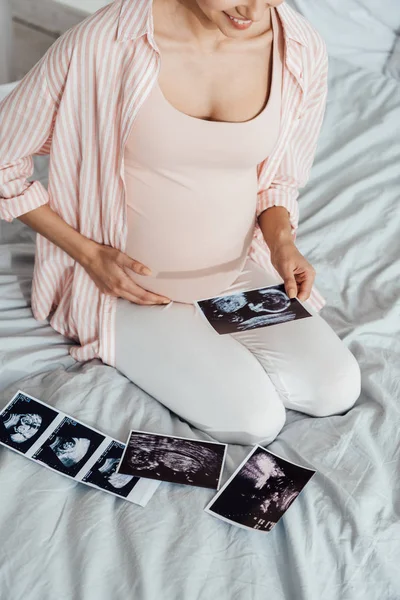 Частковий вигляд вагітної жінки, що сидить на ліжку з ультразвуковими фотографіями — стокове фото