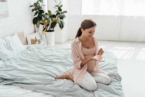 Улыбающаяся беременная женщина сидит на кровати и держит блюдце с кексом — стоковое фото