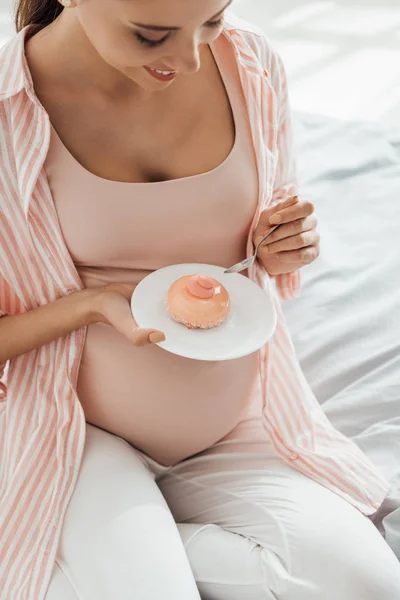 Donna incinta sorridente seduta sul letto e che tiene il piattino con cupcake — Foto stock