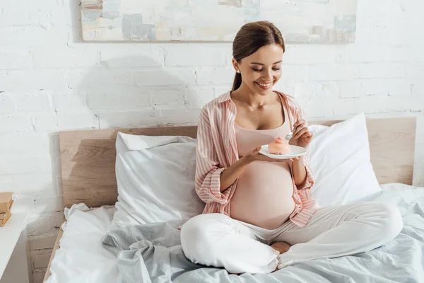 Улыбающаяся беременная женщина сидит на кровати и держит блюдце с кексом — стоковое фото