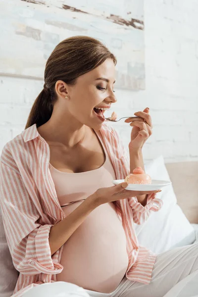 Беременная женщина сидит на кровати и ест кексы — стоковое фото