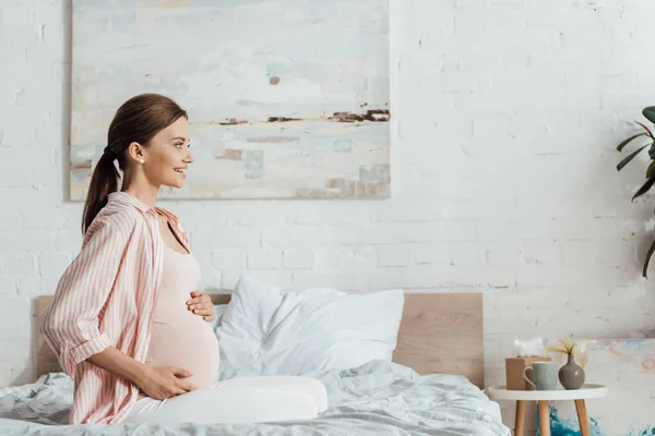 Side view of happy pregnant woman sitting on bed and touching belly — Stock Photo