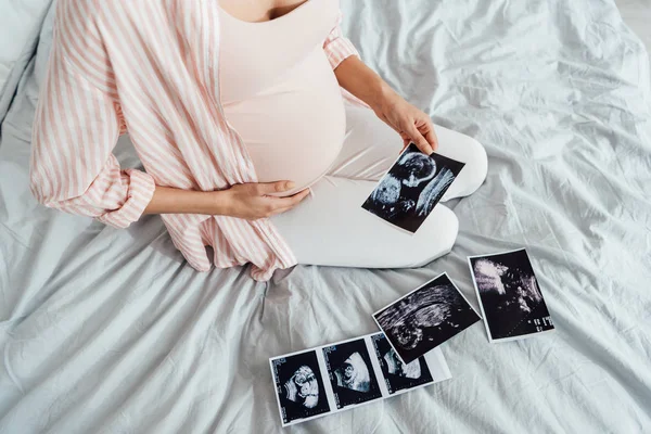 Partial view of pregnant woman sitting on bed with ultrasound pictures — Stock Photo