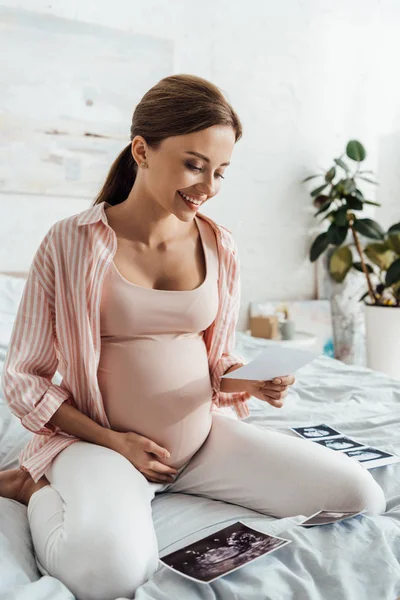 Mujer embarazada sonriente sentada en la cama y mirando la imagen de ultrasonido - foto de stock