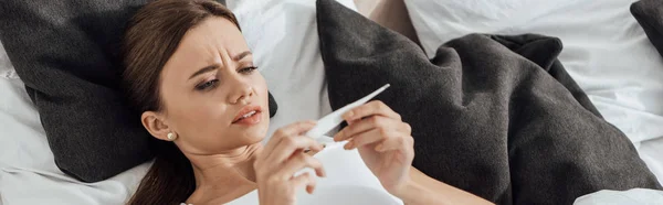 Panoramic shot of worried woman lying in bed and looking at pregnancy test — Stock Photo