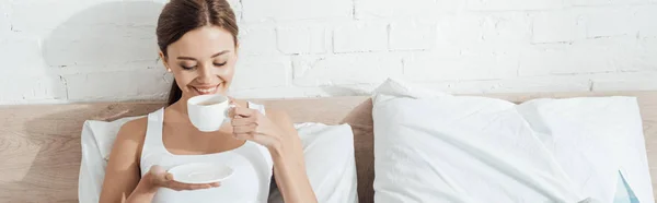 Panoramic shot of smiling woman lying in bed and drinking coffee — Stock Photo