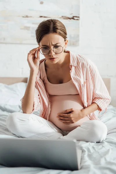 Worried pregnant woman in glasses sitting on bed and using laptop — Stock Photo