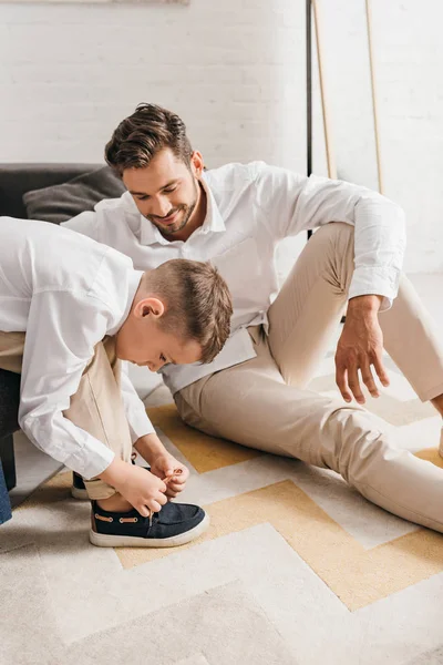 Улыбающийся отец учит сына завязывать шнурки дома — стоковое фото