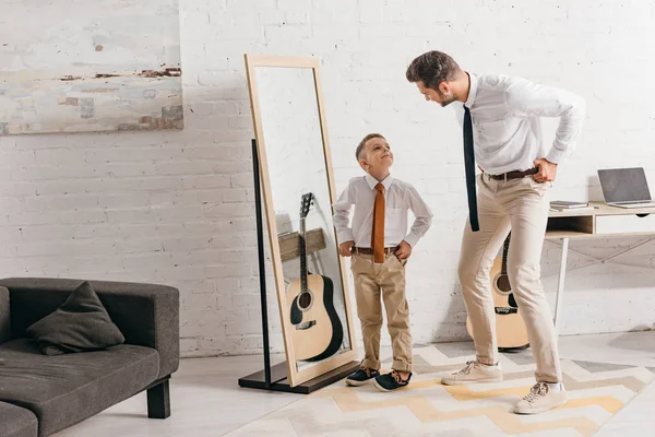 Vista completa de hijo y padre en desgaste formal de pie cerca del espejo y mirándose el uno al otro - foto de stock