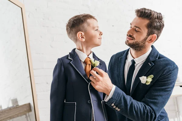 Smiling dad and son in jackets with boutonnieres looking at each other — Stock Photo
