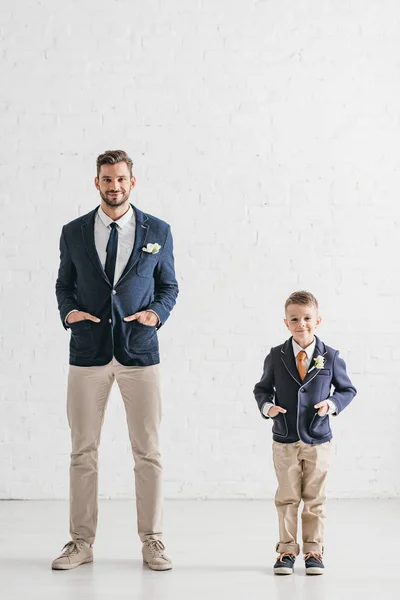 Полный вид на отца и сына в куртках с бутоньерками, стоящими с руками в карманах — стоковое фото
