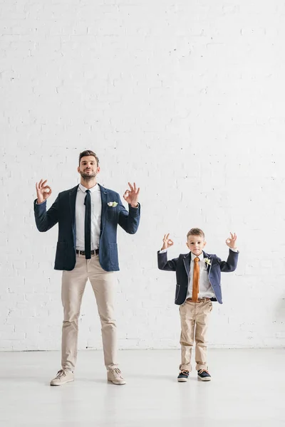 Visão de comprimento total de pai e filho em jaquetas com boutonnieres mostrando sinais ok — Fotografia de Stock