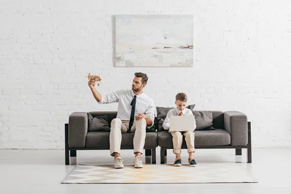 Папа с игрушечным самолетом и сын с ноутбуком сидит на диване — стоковое фото