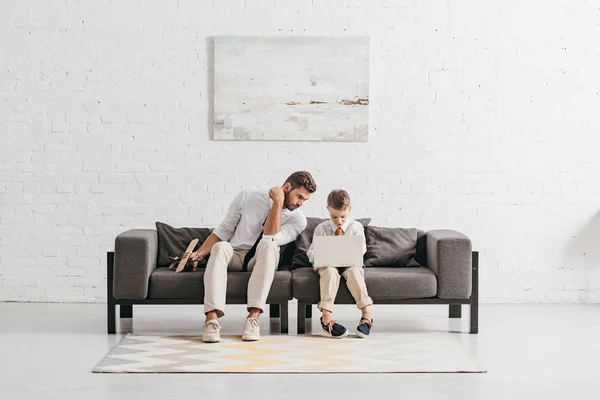 Papá con avión de juguete e hijo con portátil sentado en el sofá - foto de stock