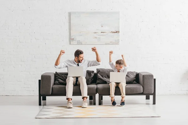 Padre e hijo en ropa formal usando computadoras portátiles y mostrando gestos de sí mientras están sentados en el sofá - foto de stock