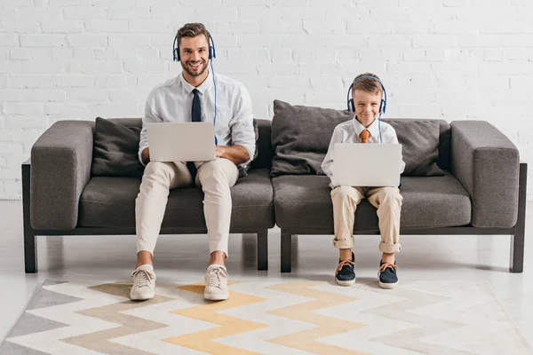 Padre e hijo en ropa formal usando computadoras portátiles y escuchando música en los auriculares mientras están sentados en el sofá - foto de stock