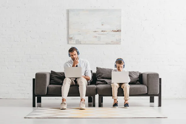 Dad and son in formal wear using laptops and listening music in headphones while sitting on sofa — Stock Photo
