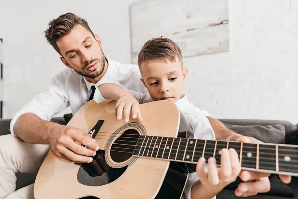 Padre enseñando hijo a tocar la guitarra acústica en casa - foto de stock