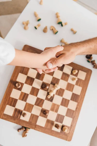 Vista parcial de papá y su hijo dando la mano sobre el tablero de ajedrez - foto de stock