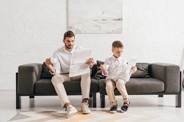 Hijo y papá en ropa formal sentado en el sofá y leyendo periódicos - foto de stock