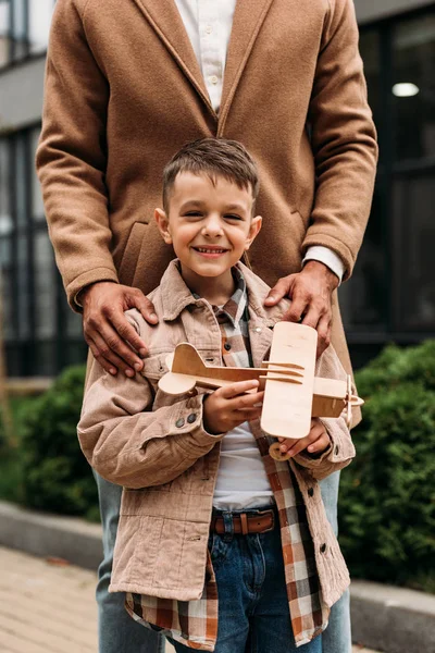 Частичный вид отца в стильном пальто и улыбающийся сын, держащий игрушечный самолет на улице — стоковое фото