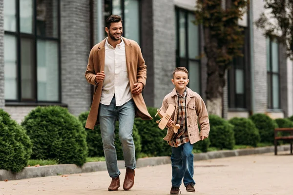 Вид в полный рост отца в пальто и сына с игрушечным самолетом, бегущим по улице — стоковое фото
