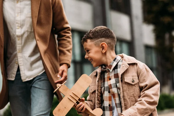 Vista parcial de papá en abrigo y niño sonriente jugando con avión de juguete de madera en la calle - foto de stock