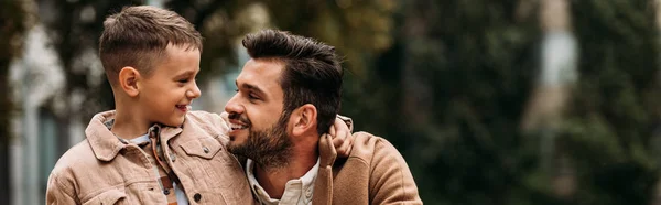 Panoramic shot of smiling son and dad embracing and looking at each other in autumn day on street — Stock Photo