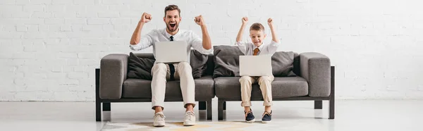 Plano panorámico de papá sonriente y el hijo sentado en el sofá con ordenadores portátiles y mostrando sí gestos - foto de stock
