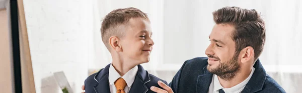 Plano panorámico de sonriente hijo y papá en ropa formal mirándose el uno al otro - foto de stock