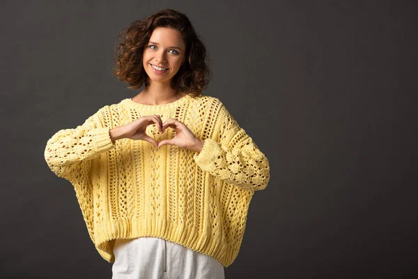 Smiling curly woman in yellow knitted sweater showing heart sign on black background — Stock Photo