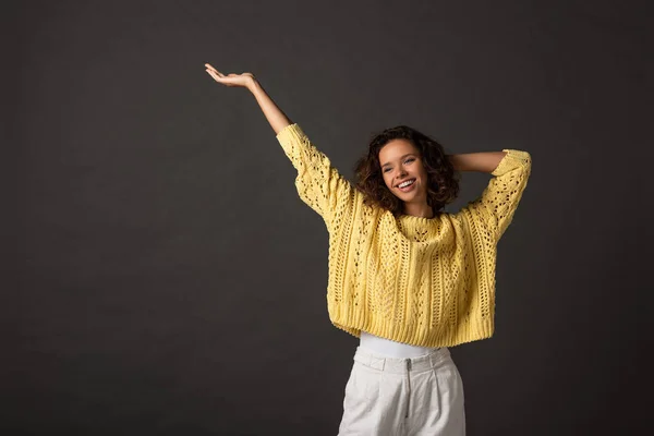 Sonriente mujer rizada en suéter de punto amarillo con las manos en el aire sobre fondo negro - foto de stock