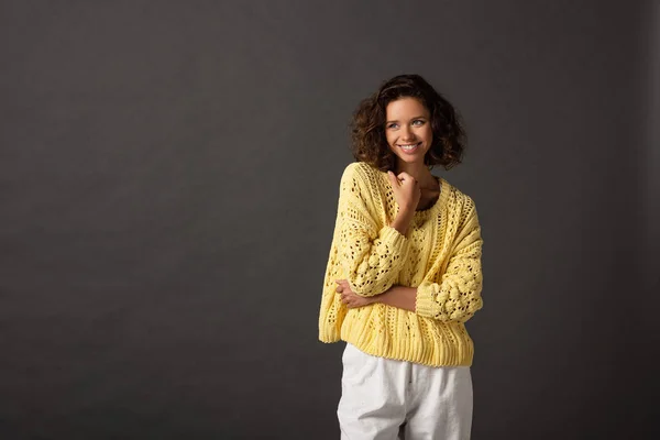Smiling curly woman in yellow knitted sweater looking away on black background — Stock Photo