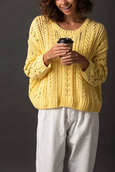 Cropped view of happy woman in yellow knitted sweater holding coffee to go on black background — Stock Photo