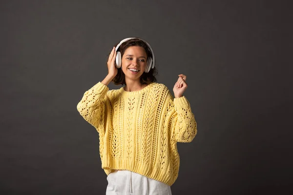 Smiling curly woman in yellow knitted sweater listening music in headphones on black background — Stock Photo