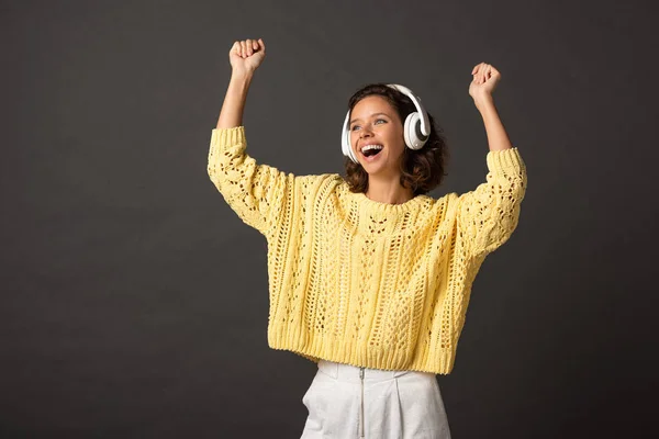 Happy curly woman in yellow knitted sweater with hands in air listening music in headphones on black background — Stock Photo