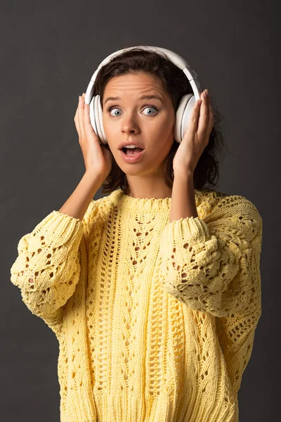 Femme bouclée choquée en chandail tricoté jaune écoutant de la musique dans des écouteurs sur fond noir — Photo de stock