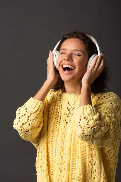 Happy curly woman with closed eyes in yellow knitted sweater listening music in headphones on black background — Stock Photo