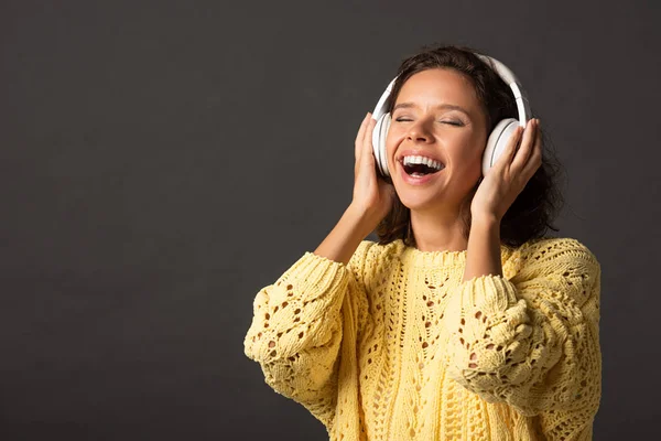 Happy curly woman with closed eyes in yellow knitted sweater listening music in headphones on black background — Stock Photo