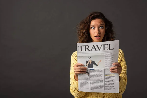 Shocked curly woman in yellow knitted sweater holding travel newspaper on black background — Stock Photo