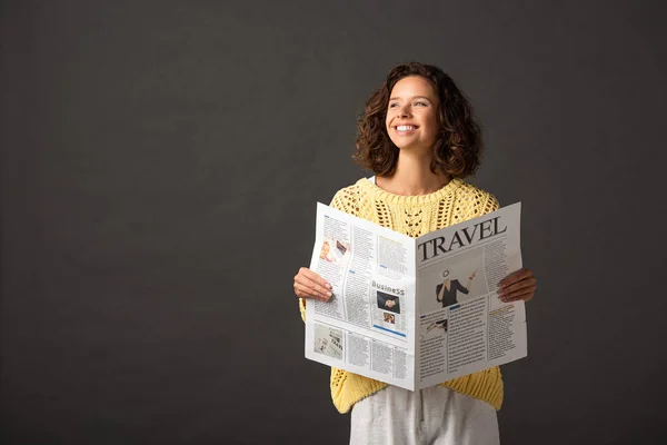 Улыбающаяся кудрявая женщина в желтом трикотажном свитере держит газету о путешествиях на черном фоне — стоковое фото