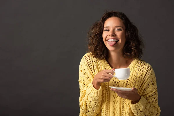 Felice donna riccia in maglia gialla maglione mostrando la lingua e tenendo la tazza di caffè e piattino su sfondo nero — Foto stock