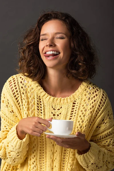 Heureuse femme bouclée en chandail tricoté jaune montrant la langue avec les yeux fermés et tenant tasse de café et soucoupe sur fond noir — Photo de stock