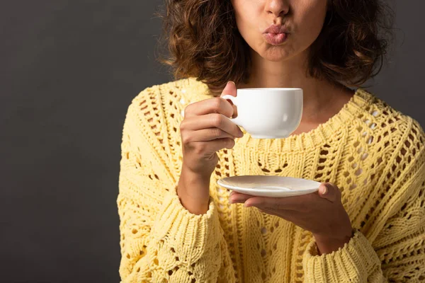Cropped view of curly woman in yellow knitted sweater pouting lips while holding cup of coffee and saucer on black background — Stock Photo