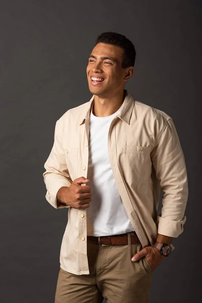 Happy handsome mixed race man in beige shirt with hand in pocket on black background — Stock Photo