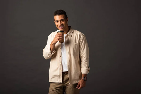 Sorrindo bonito misto homem de raça em camisa bege segurando café para ir no fundo preto — Fotografia de Stock