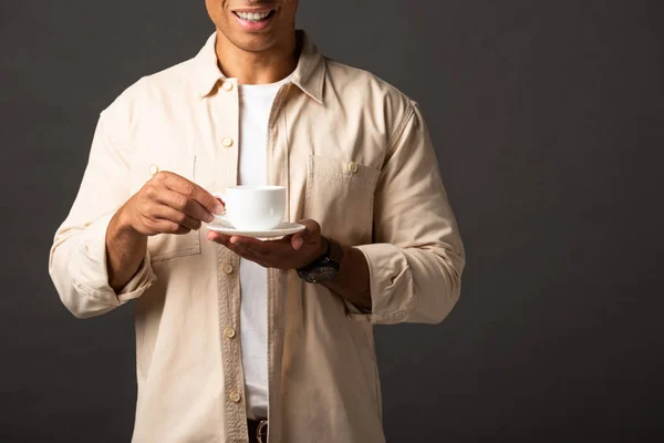 Vista recortada de sonriente hombre de raza mixta en camisa beige sosteniendo taza de café y platillo sobre fondo negro - foto de stock