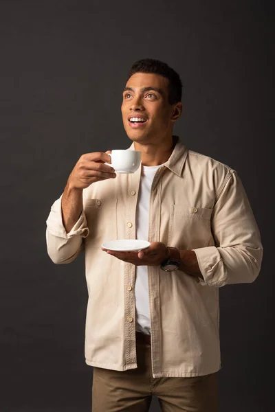 Lächelnder Mischlingsmann im beigen Hemd, der wegschaut und eine Tasse Kaffee und Untertasse auf schwarzem Hintergrund hält — Stockfoto
